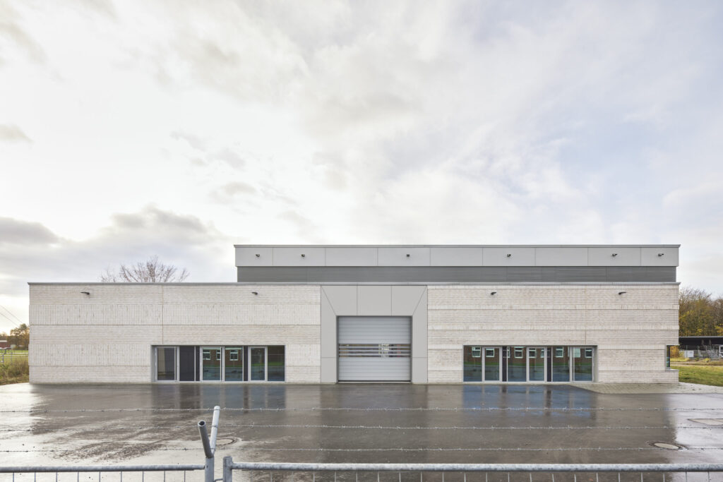 Flaches, rechteckiges Gebäude mit hellen Fassadenplatten, großen Fenstern und zentraler Toröffnung, davor nasser Asphalt.