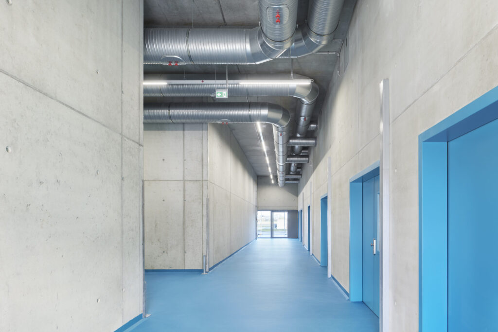 Innenraum mit blauen Türen und blauen Bodenbelag, graue Betonwände und sichtbare Lüftungsrohre an der Decke.