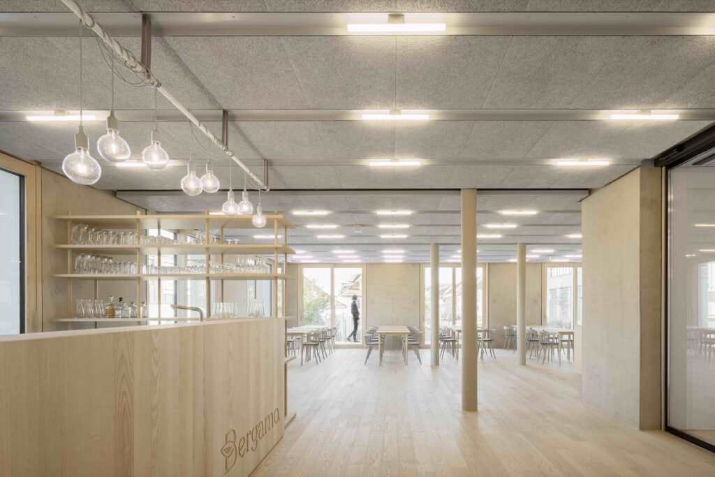 Heller, moderner Gastraum mit Holzfußboden, Holzsäulen, Holztheke mit Schriftzug 'Bergamo' und mehreren Tischen und Stühlen.