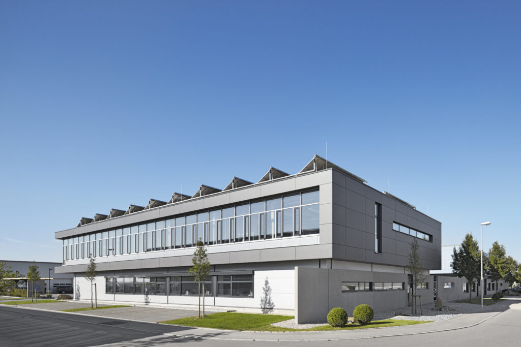 Modernes, zweistöckiges Bürogebäude mit großen Fenstern, grauer Fassade und kleinen Bäumen davor.