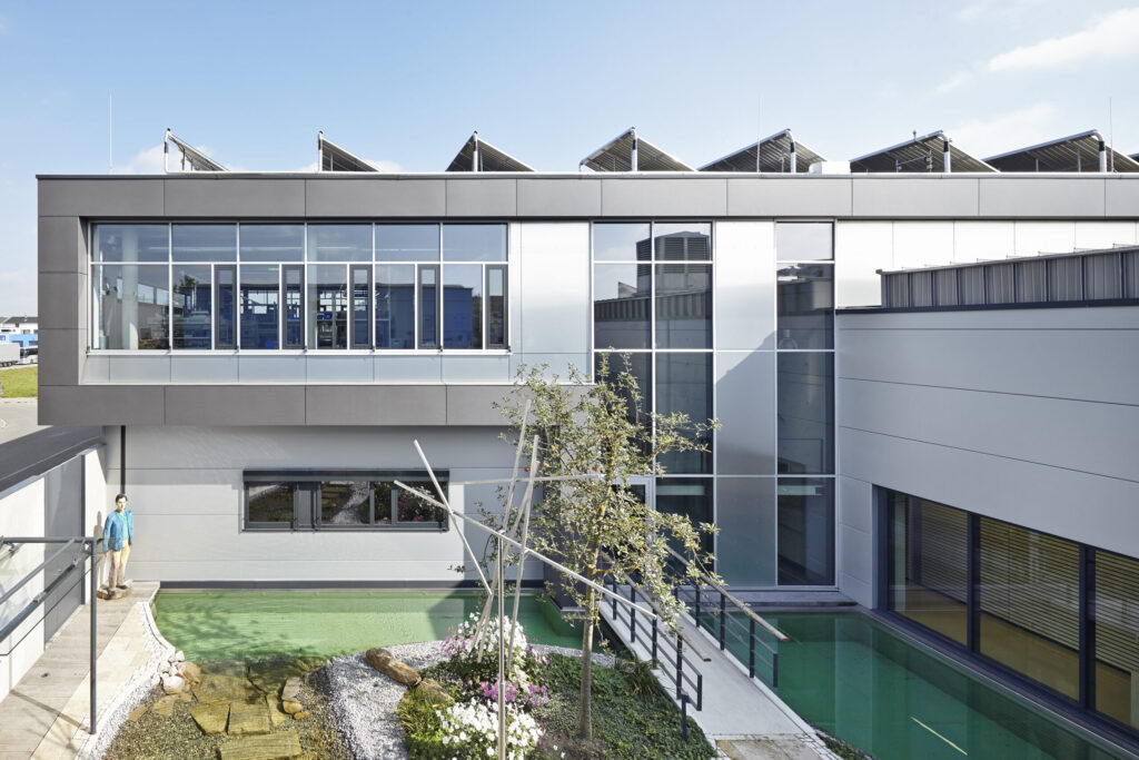 Modernes Gebäude mit großen Fenstern, Solarpaneelen auf dem Dach und einem kleinen Garten mit Baum und Wasserbecken im Innenhof.