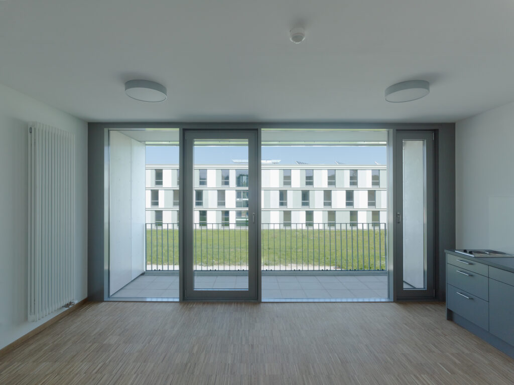 Leerer Raum mit Parkettboden, großer Glasfront mit drei Türen und Blick auf ein modernes Wohngebäude mit weißer Fassade und Balkonen.