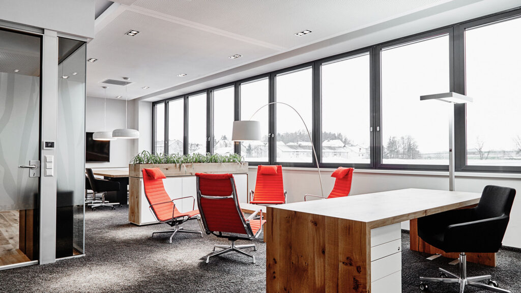 Modernes Büro mit großen Fenstern, Holztisch, mehreren roten und einem schwarzen Bürostuhl, Teppichboden und Stehlampen.