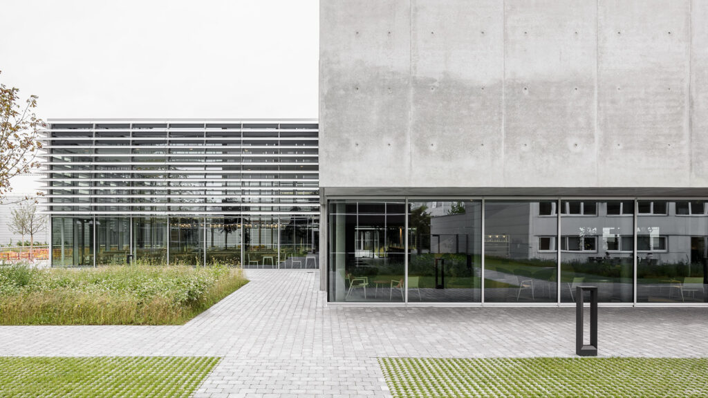 Modernes Gebäude mit großflächigen Glasfronten und Sichtbetonfassade, davor gepflasterter Platz und Grünflächen.
