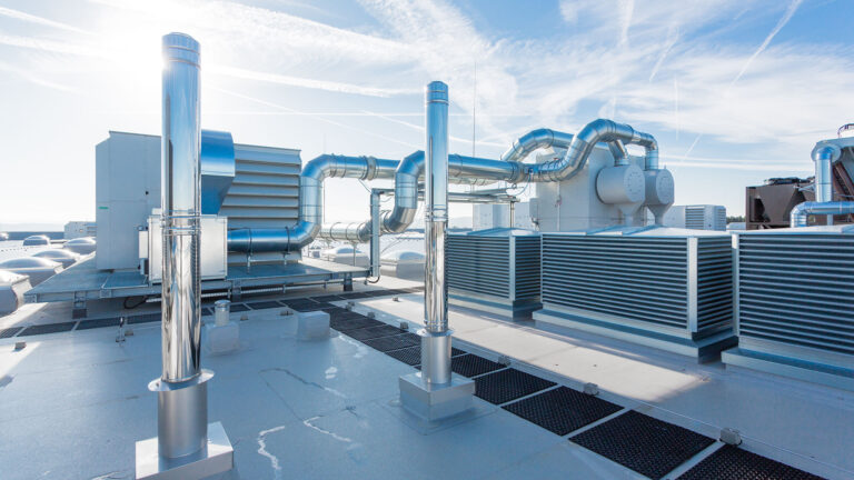 Dach mit mehreren silbernen Lüftungsrohren und großen grauen Lüftungsanlagen unter blauem Himmel.